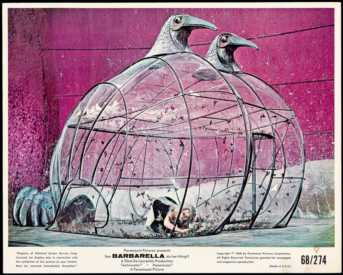 A woman is seen inside a large, transparent cage topped with two giant bird heads, set against a purple background with scattered feathers. Zoom in to explore the details of this Movie Still (Paramount 1968).